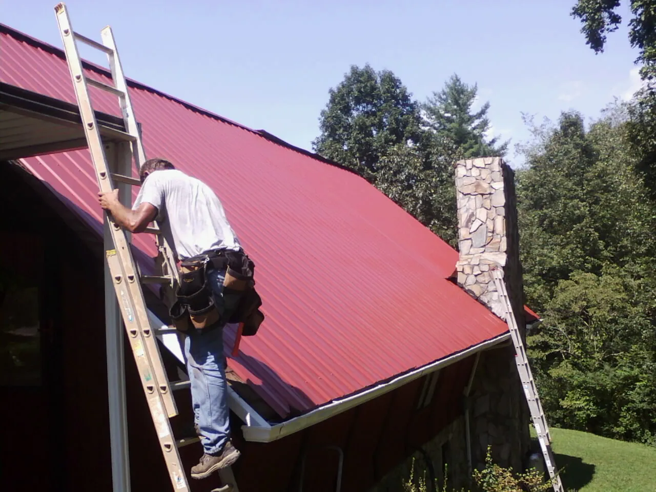 Roofer on a ladder beside a red metal roof and stone chimney — Soffit & Fascia Repair by Roofing Brothers in Muldoon
